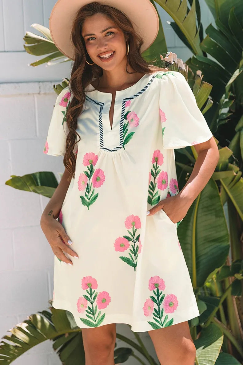 Woman wearing a white dress with pink floral embroidery in front of green foliage.