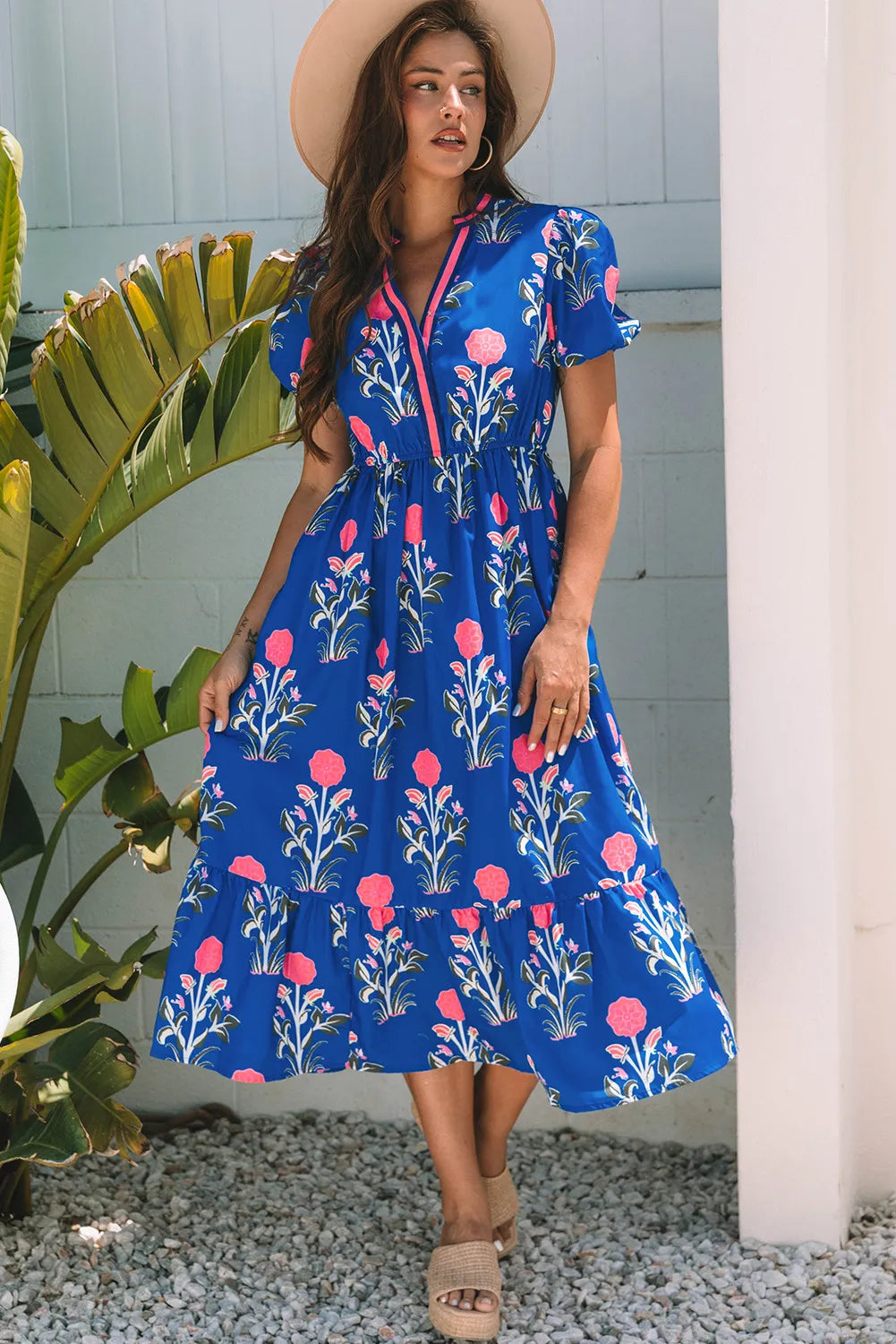 Woman wearing a blue floral dress standing outdoors with plants and a column in the background.