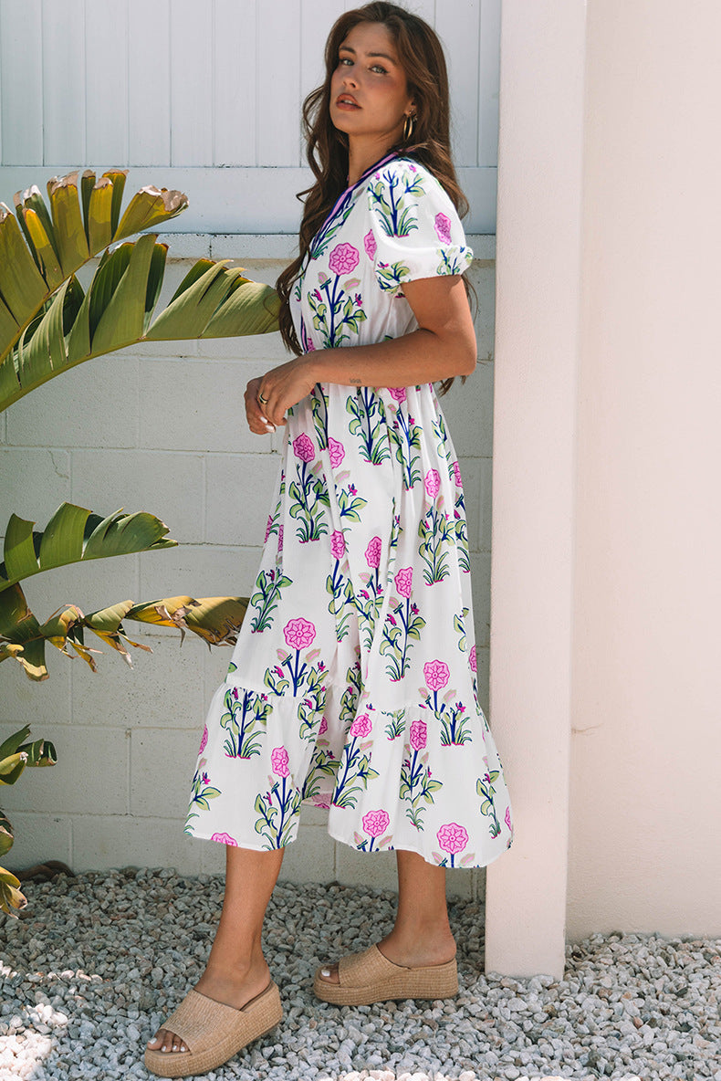 Woman wearing a floral dress standing outdoors with plants and a white wall in the background