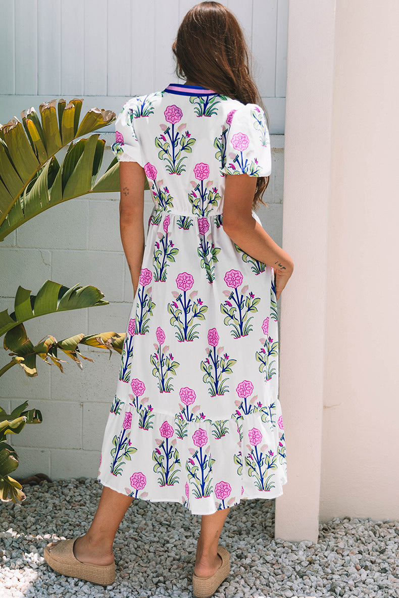Woman wearing a floral dress standing outdoors with plants and a white wall in the background