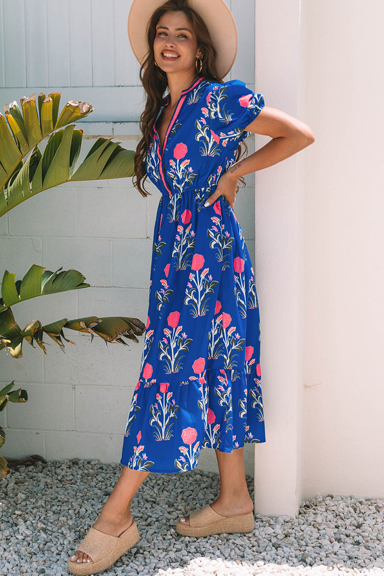 Woman wearing a blue floral dress standing against a white wall with greenery.