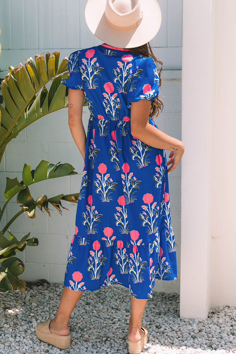 Woman wearing a blue floral dress standing outdoors with greenery and a white column in the background.